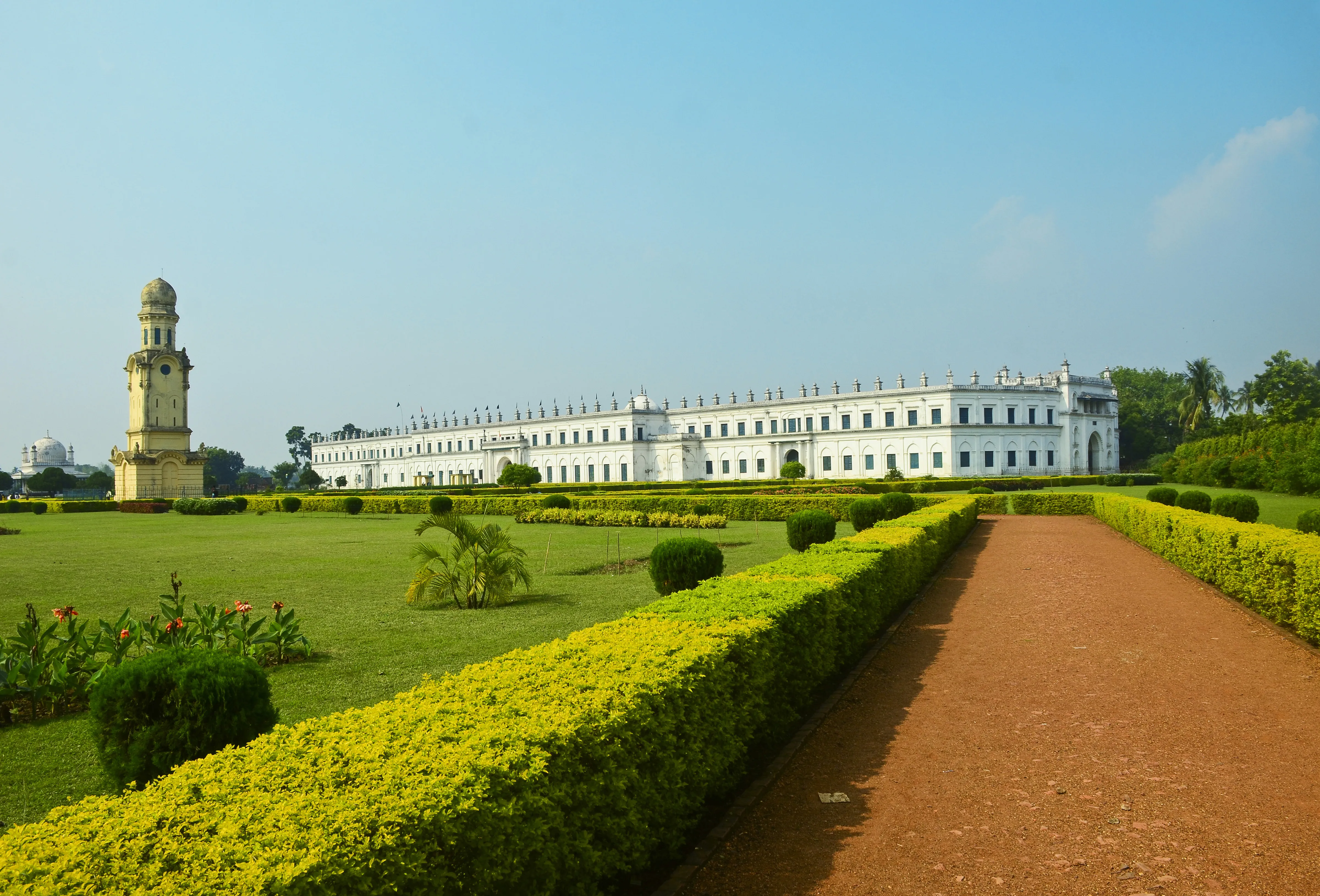 A long white colonnaded building with an arched gateway and rows of small finials along its roofline, seen across a formal lawn with clipped hedges, a domed building and a clock tower visible to the left