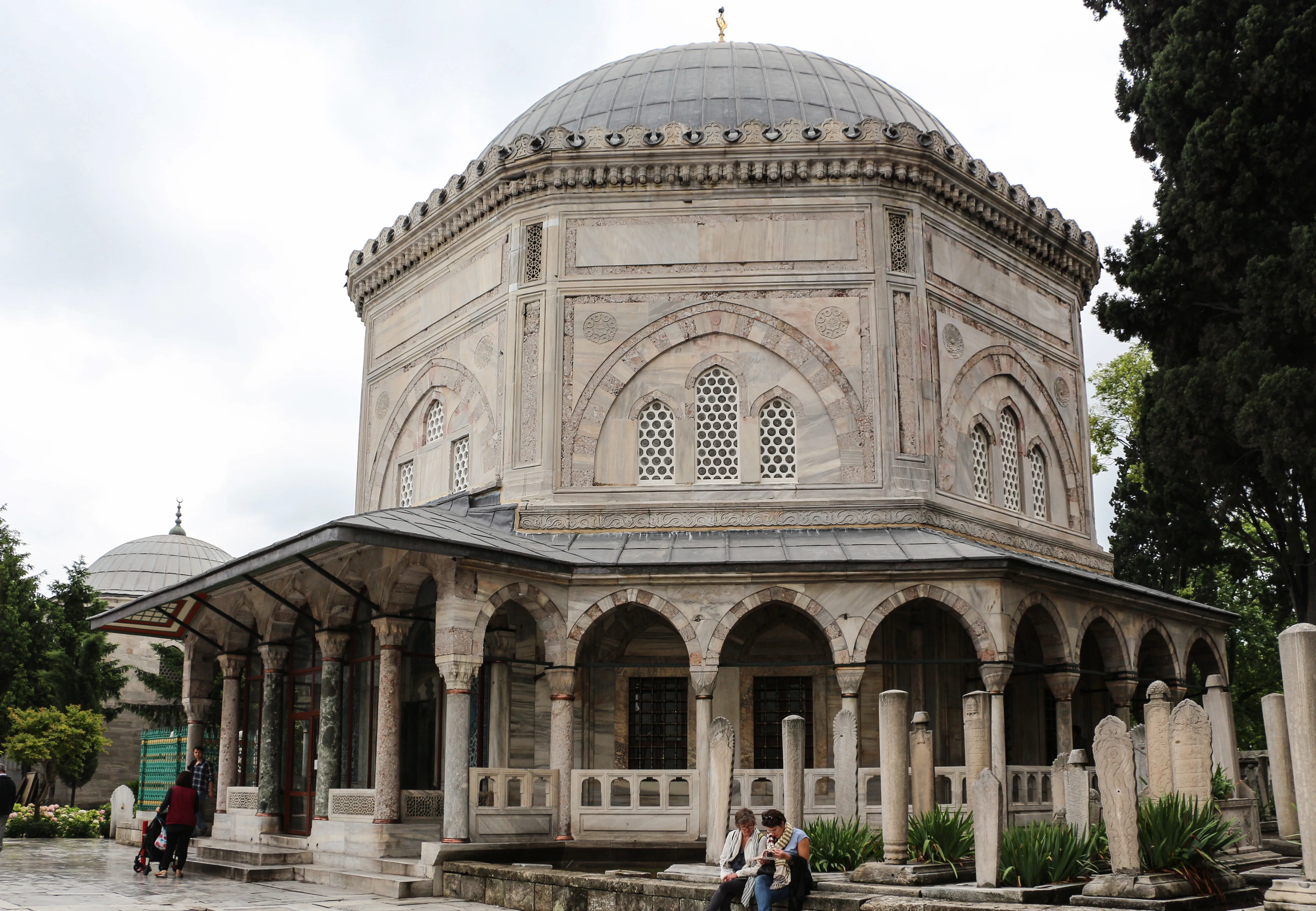 The interior of a domed mausoleum with a large cloth-draped cenotaph topped by a wrapped turban, gold Arabic calligraphy on the covering, patterned tile walls, arched stained-glass windows, and a large crystal chandelier overhead