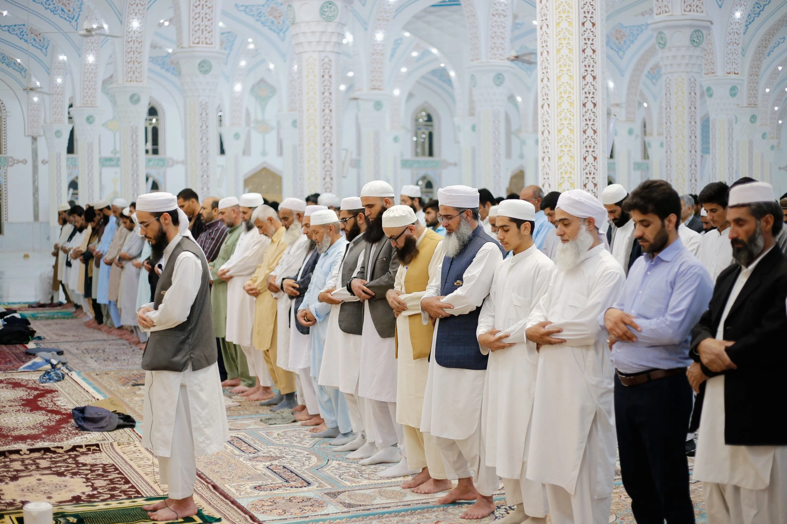 Rows of men standing in congregational prayer with hands folded, on patterned carpets inside a mosque with white columns and arches decorated in blue and turquoise