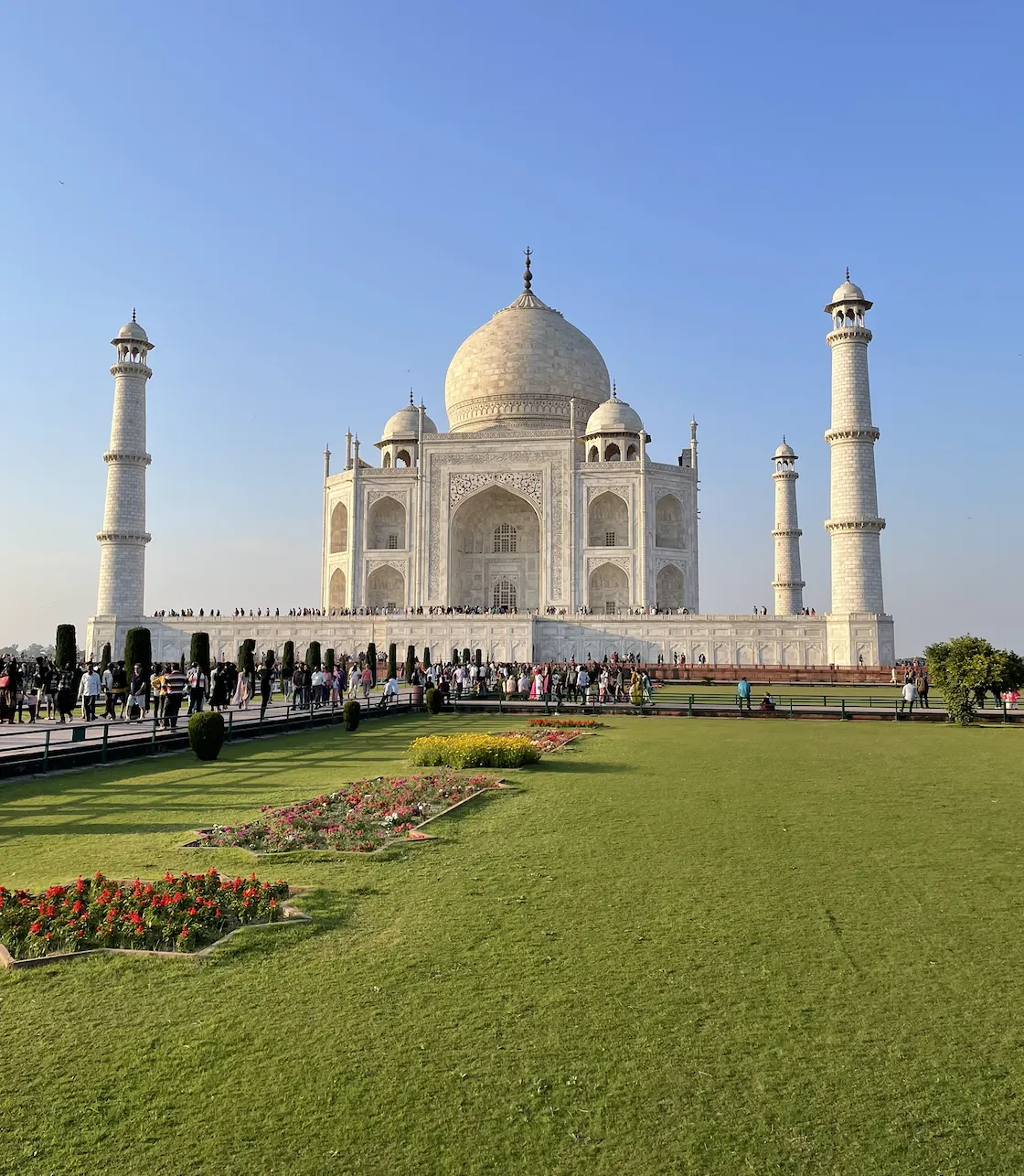 The Taj Mahal in white marble with its central dome and four flanking minarets, seen across the formal garden with flowerbeds and lawns under a clear sky