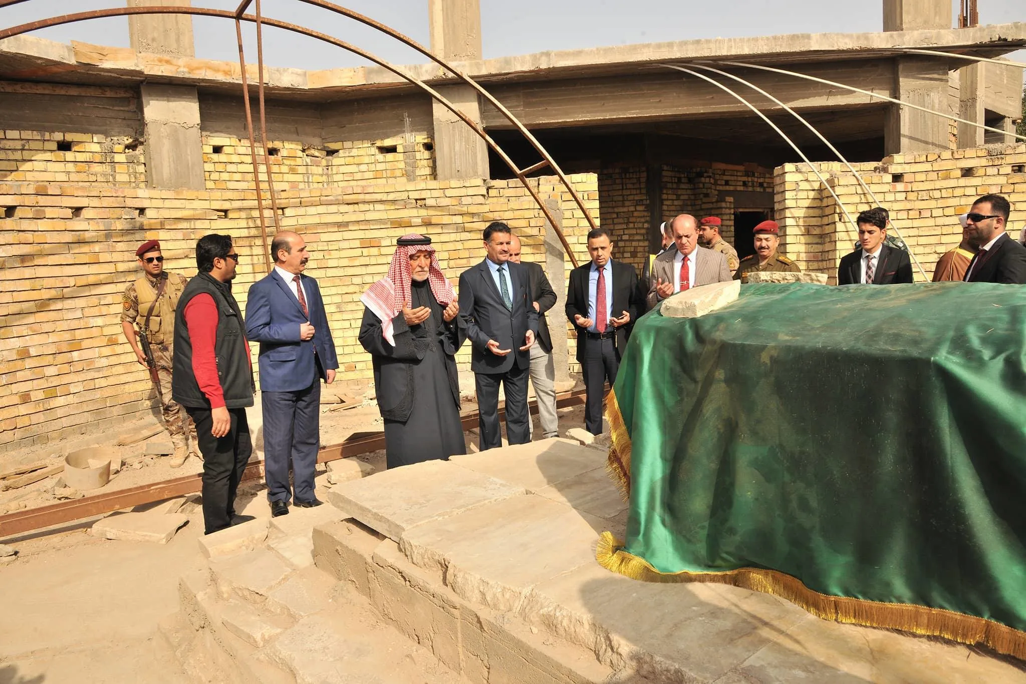 A grave draped in green cloth with a gold fringe on a raised stone base, with men in suits, an elderly man in a red-and-white keffiyeh and several uniformed soldiers standing around it, some with hands raised in supplication, before an unfinished concrete and yellow-brick building