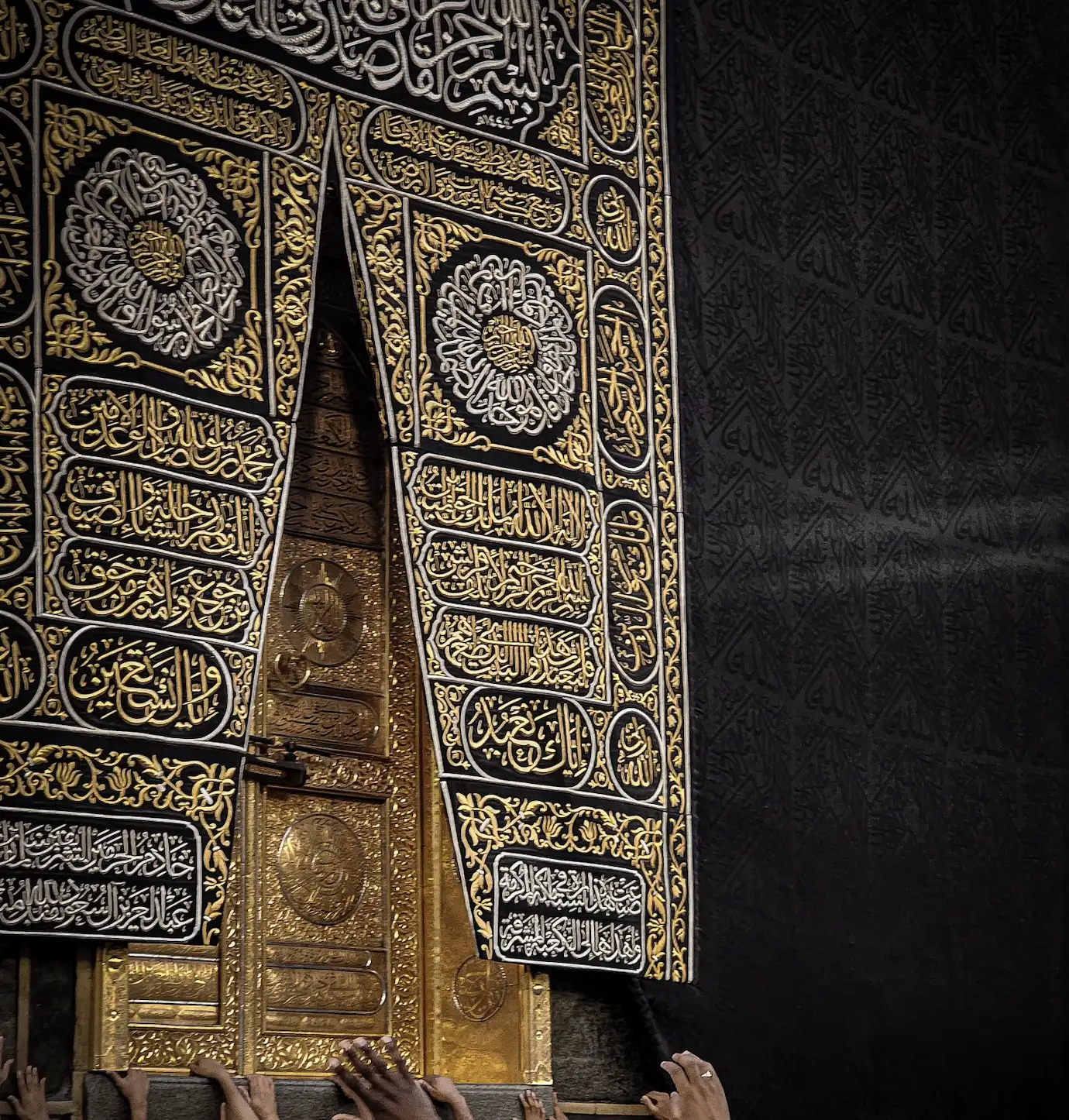 The door of the Kaaba, gold-coloured with embossed roundels, partly covered by its black curtain embroidered in gold and silver with Arabic calligraphy in panels, cartouches and two large roundels, beside a black wall covering woven with repeated calligraphy; outstretched hands reach up to the threshold at the bottom of the frame.