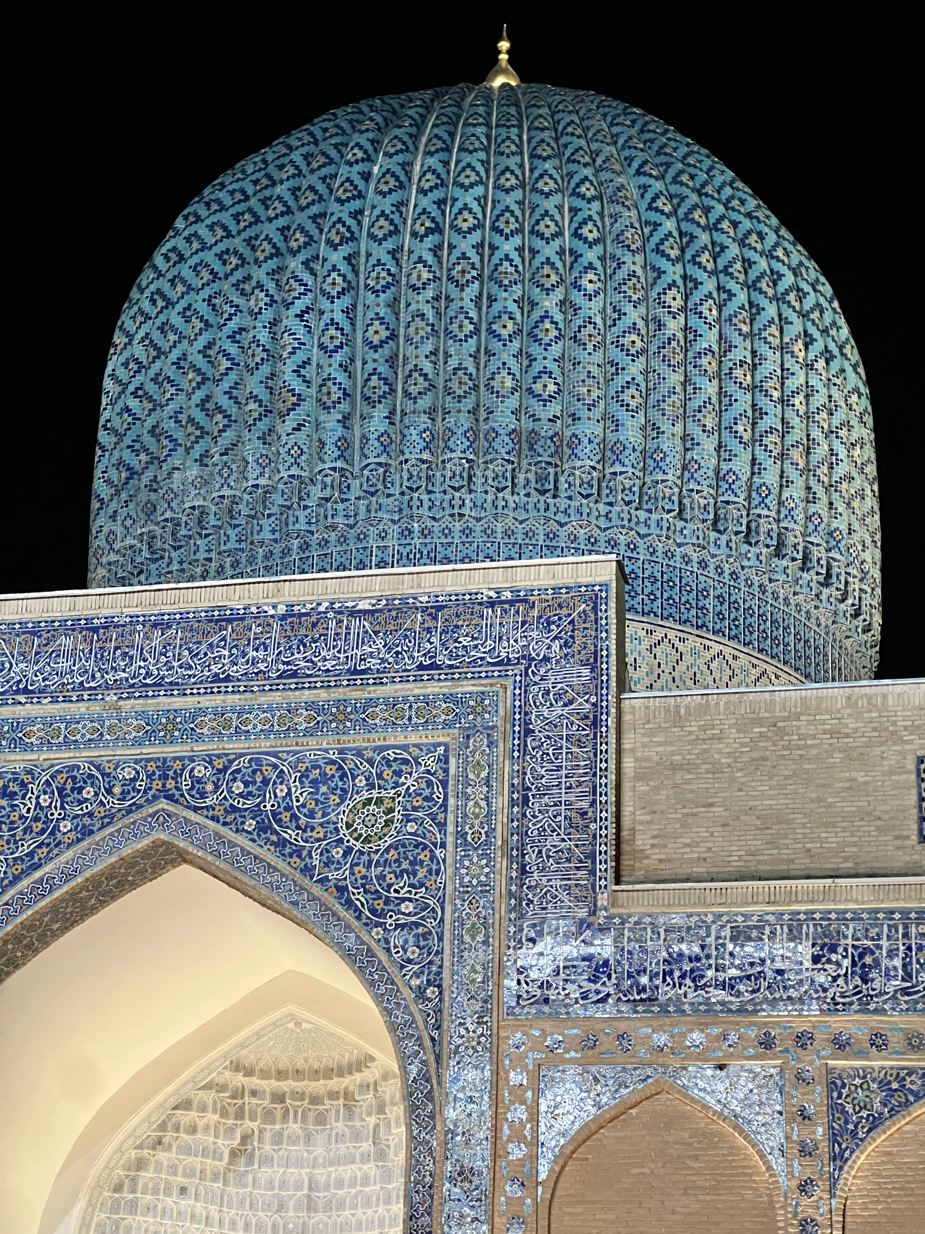 A close night view of a ribbed turquoise-tiled dome patterned with diamond motifs, rising above the tiled and mirror-inlaid corner of an iwan carrying bands of calligraphy, seen against a black sky