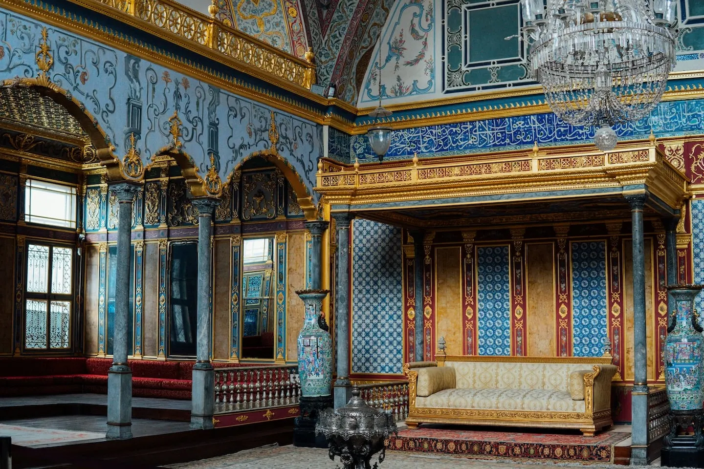 A domed interior at Topkapi Palace: a canopied dais carrying a gilded couch, set against panels of blue and white tilework framed in gold and marble, with painted blue and gold wall decoration, a band of calligraphy in blue and white running above, marble columns, large enamelled vases, a silver brazier and a crystal chandelier