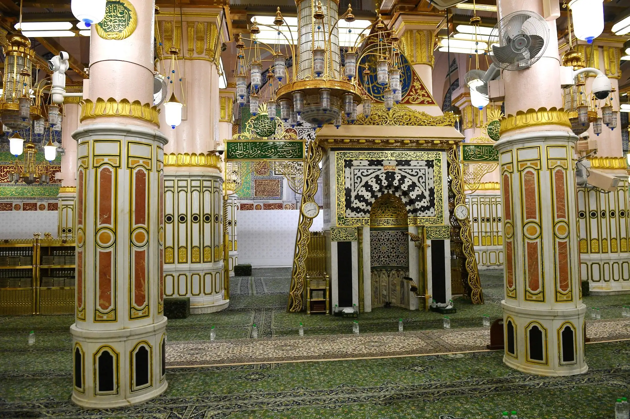 A columned interior of a mosque richly decorated in gold, white marble and green: a mihrab niche framed in black-and-white geometric inlay and gold calligraphic panels stands beside a marble column with gold-crowned capital, chandeliers and hanging lanterns above, and a green sign in gold Arabic lettering marking the Rawdah, with worshippers' bottled water set along the patterned green carpet