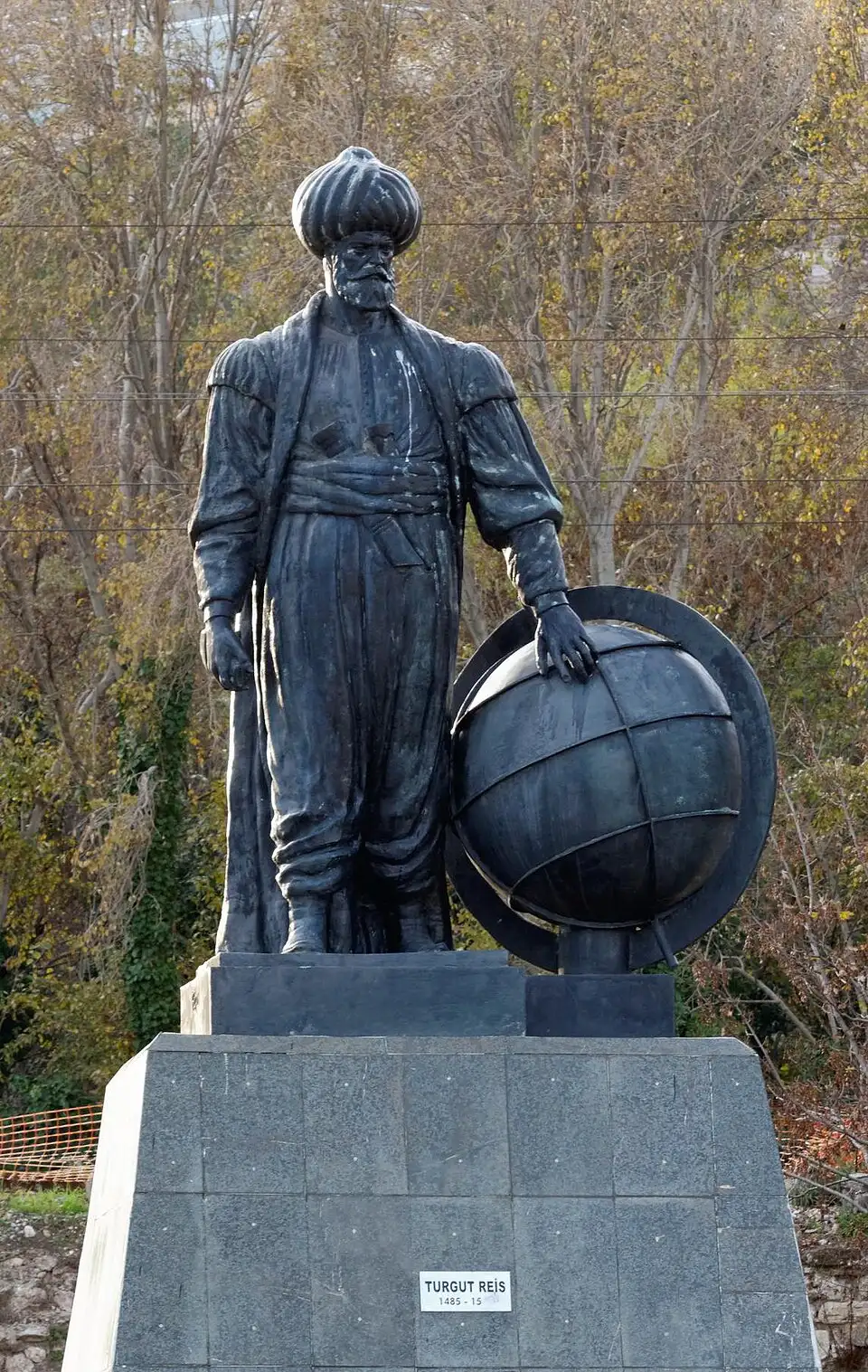 A dark metal statue of a bearded man in a tall turban, long caftan and sash, standing with one hand resting on a large globe, on a stone plinth bearing a small plaque with his name, with autumn trees behind