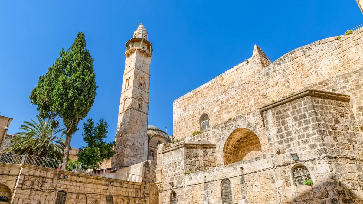 The stone minaret and domed building of the Mosque of Omar in the Old City of Jerusalem, beside the ancient stone walls of the Church of the Holy Sepulchre, with a cypress and palm tree against a blue sky