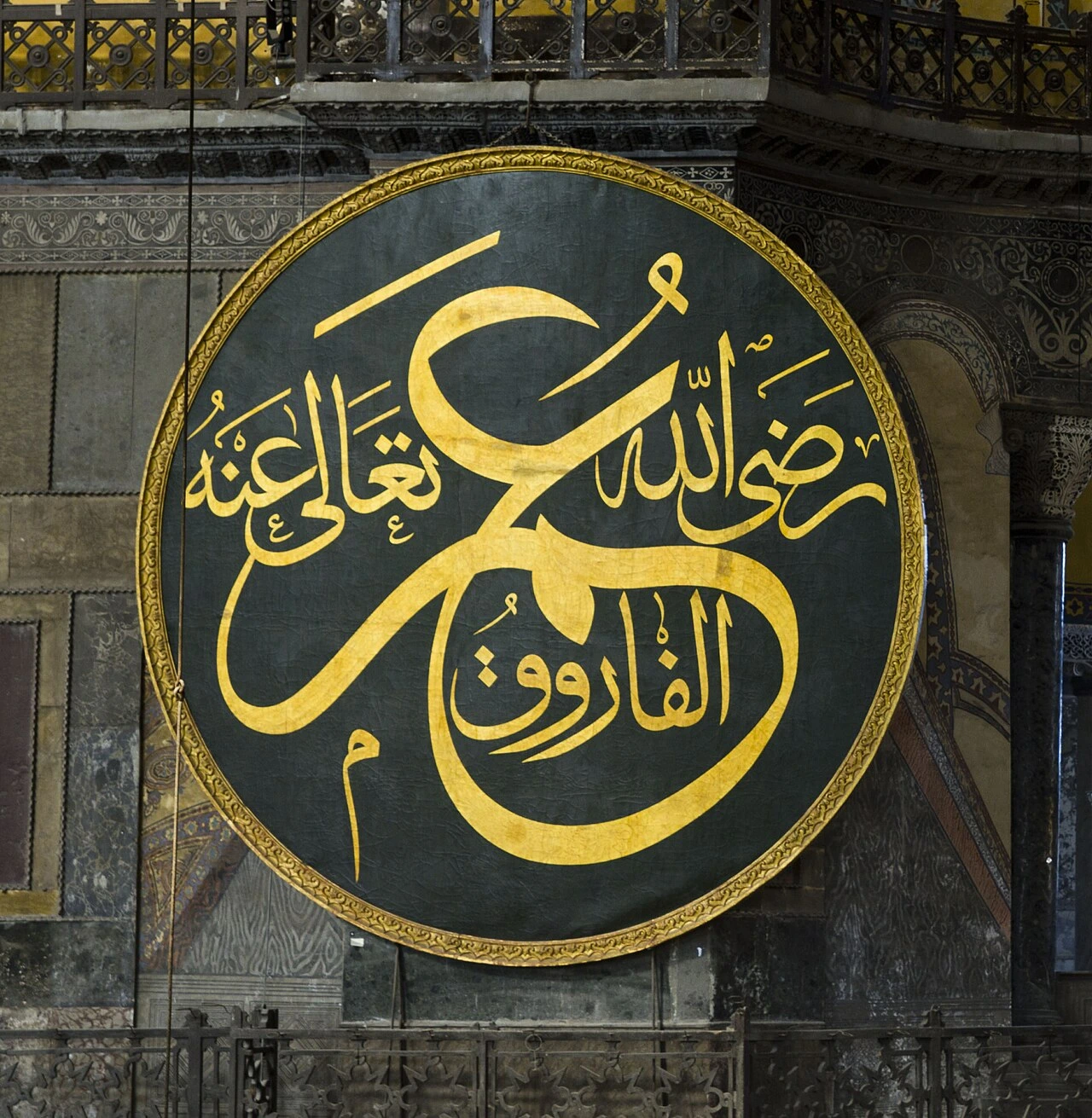 A large circular calligraphic medallion in gold script on a dark green ground, framed in gilt and mounted on a marble-clad wall beneath a gallery, giving the name Umar with the epithet al-Faruq
