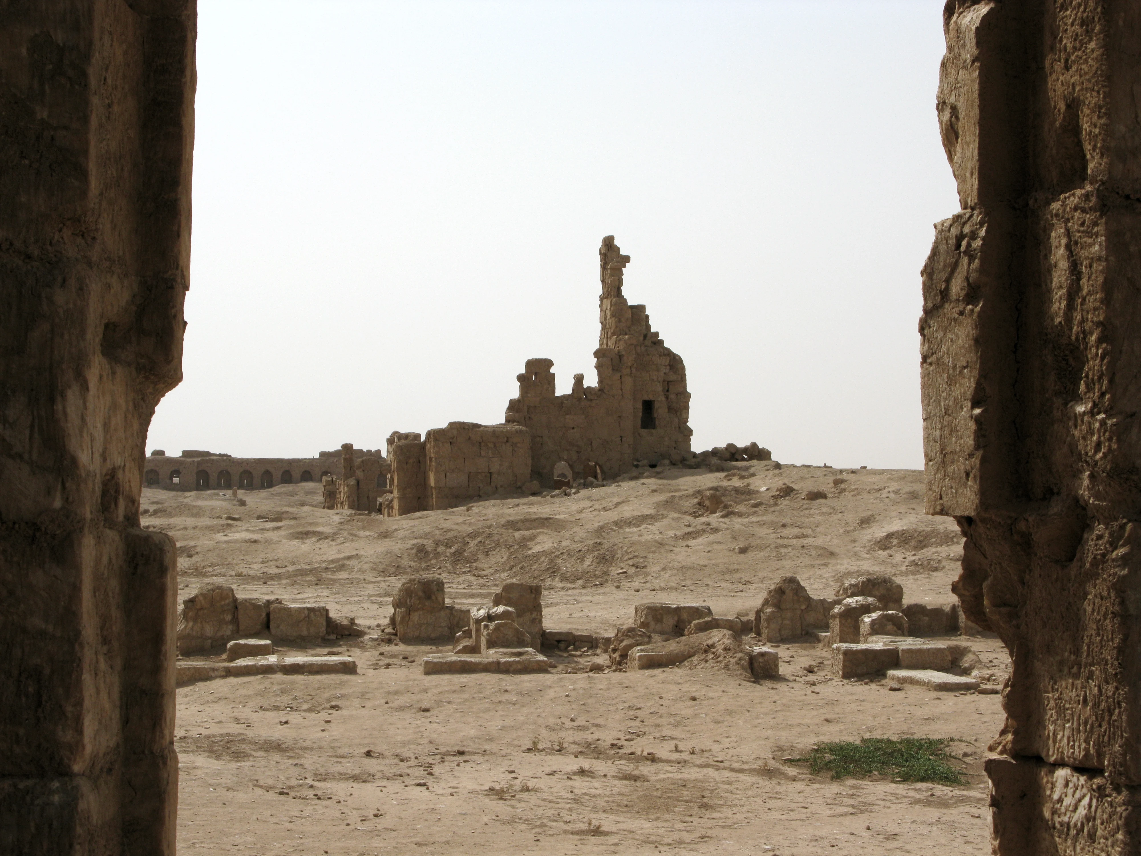 Ruined stone walls and the broken stump of a tower in a desert city, seen through a gap between two massive weathered stone blocks, with fallen masonry scattered across the sandy ground and a long arcaded wall in the distance