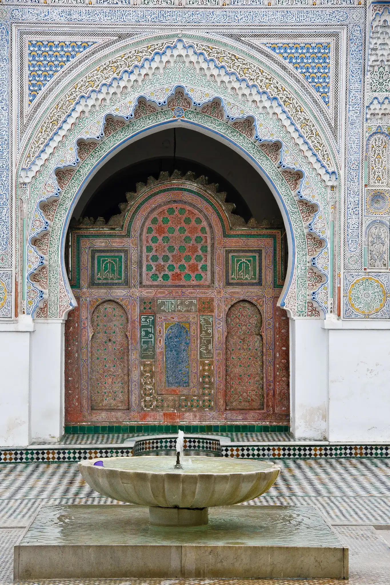 A large ornately carved and painted horseshoe arch at the al-Qarawiyyin mosque in Fez, covered in stucco, Arabic inscription bands and coloured zellij tilework, with a scalloped marble fountain on a checkerboard-tiled floor in the foreground