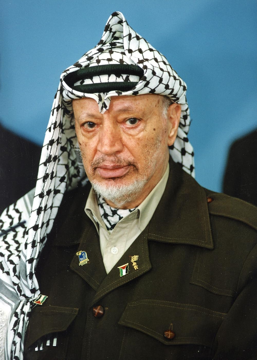 An elderly man in a black-and-white chequered keffiyeh held by a dark cord, wearing an olive-green field jacket with breast pockets over a light olive shirt, with a small Palestinian flag pin on the lapel, against a plain blue background