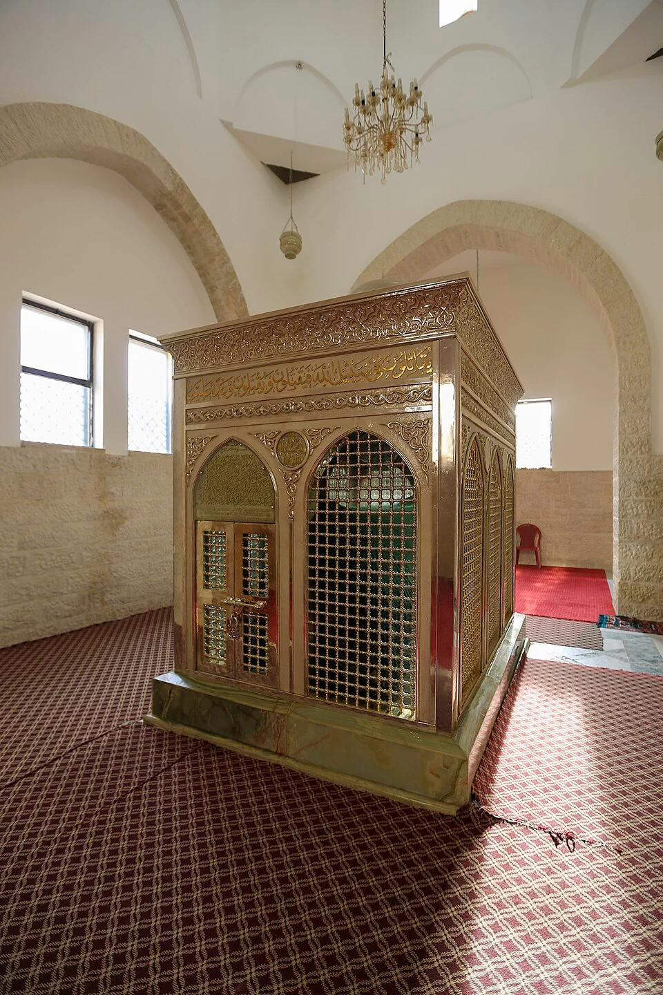 Interior of a shrine with a gilded cenotaph enclosure decorated with Arabic calligraphy and latticework, standing under whitewashed vaulted arches on a patterned red carpet