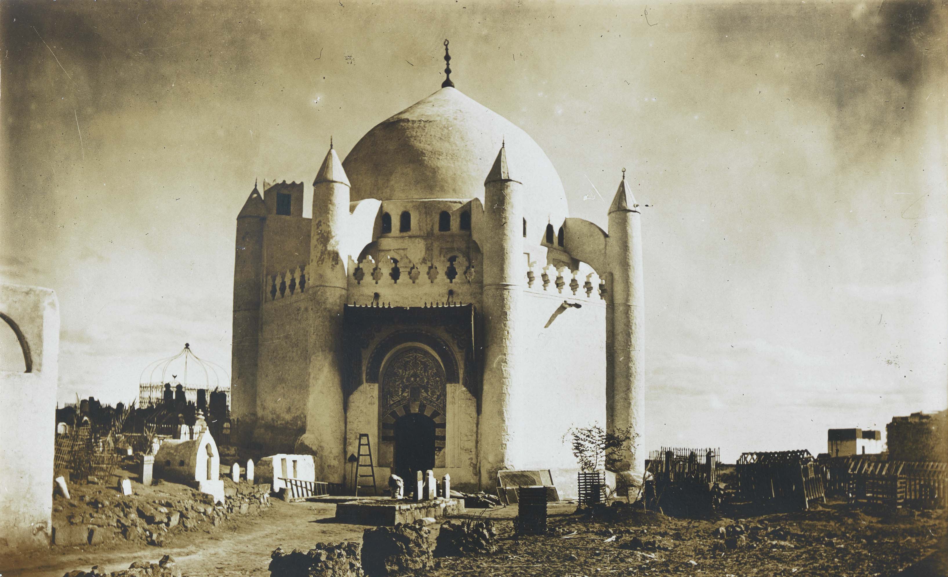 An early sepia photograph of a large whitewashed mausoleum with a tall bulbous dome, four slender corner turrets and a deeply recessed carved portal, standing on open stony ground among low grave markers and wooden railings