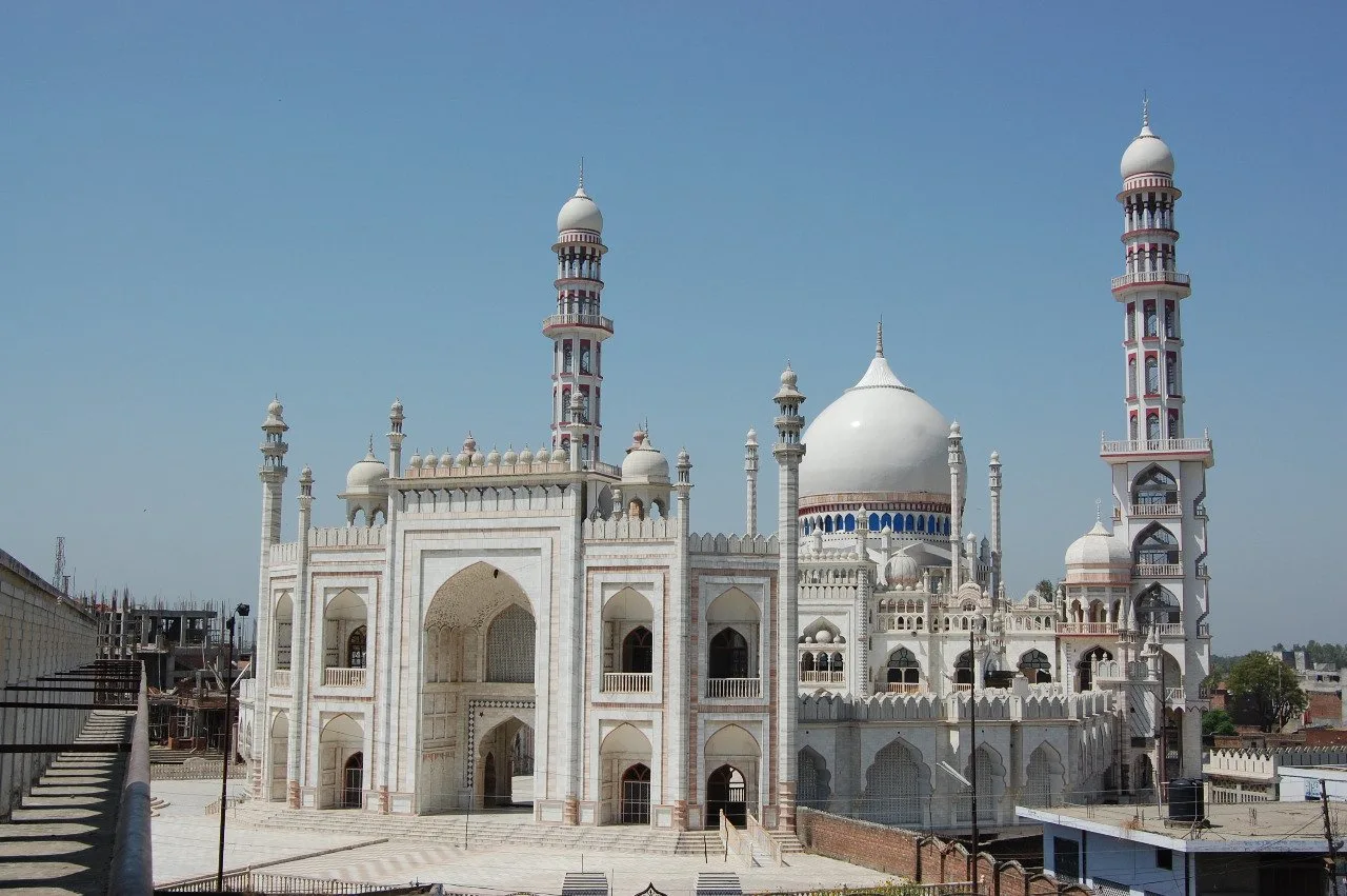 A white marble mosque complex with a large central dome, a monumental arched entrance portal, small domed pavilions along the roofline and tall slender minarets banded in red, seen from an elevated viewpoint against a clear sky